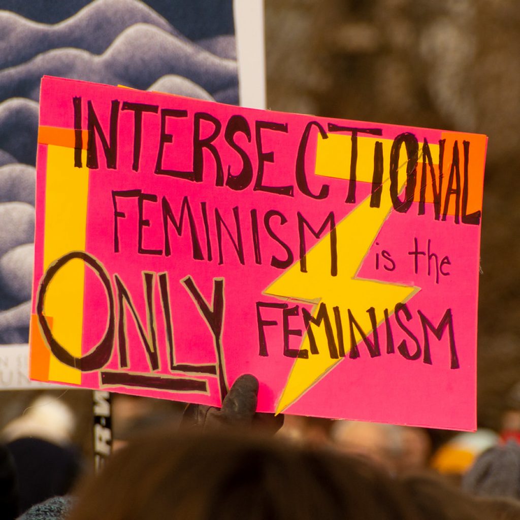 Hand written sign reading "intersectional feminism is the only feminism", black text on pink background with yellow pattern. The sign is the only thing in focus, but below it the tops of marcher's heads are visible out of focus in front of and behind the sign.