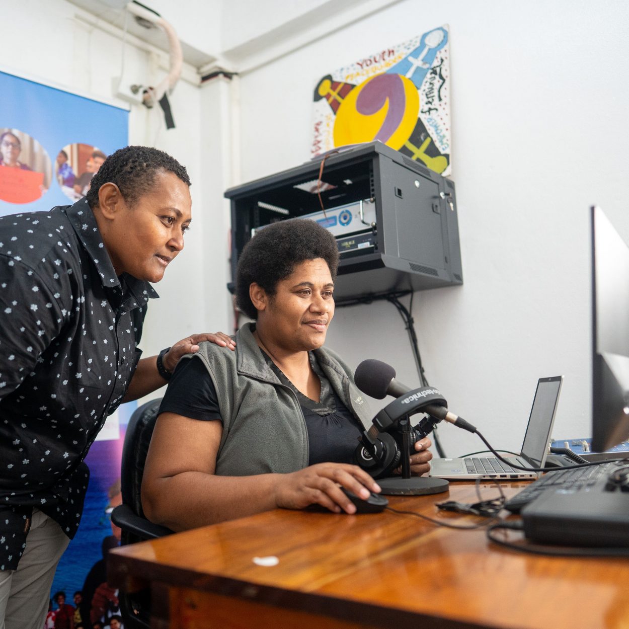 Photo: One woman sits at a desk with a microphone and a computer, another woman leans over her shoulder watching her work