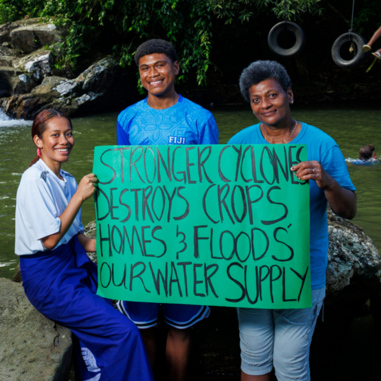 A young woman, a young man and a woman hold a sign that says Stronger cyclones destroys crops homes and floods our water supply. Behind them, is a natural swimming hole where young teenage boys play on tyre swings above the water
