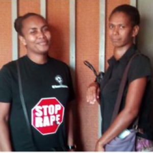 Two women from the family support centre stand in front of a wall, smiling at the camera. Both wear black shirts but the woman on the left has a "stop rape" slogan printed on her shirt in the shape of a stop sign