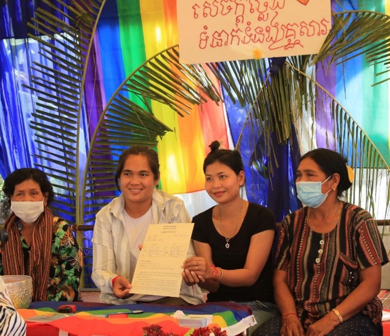 Channa Meng and Sopheap Cheam sit between two older women. They are holding a certificate. Behind them hangs a rainbow flag. A second rainbow flag covers the table that they sit at. 