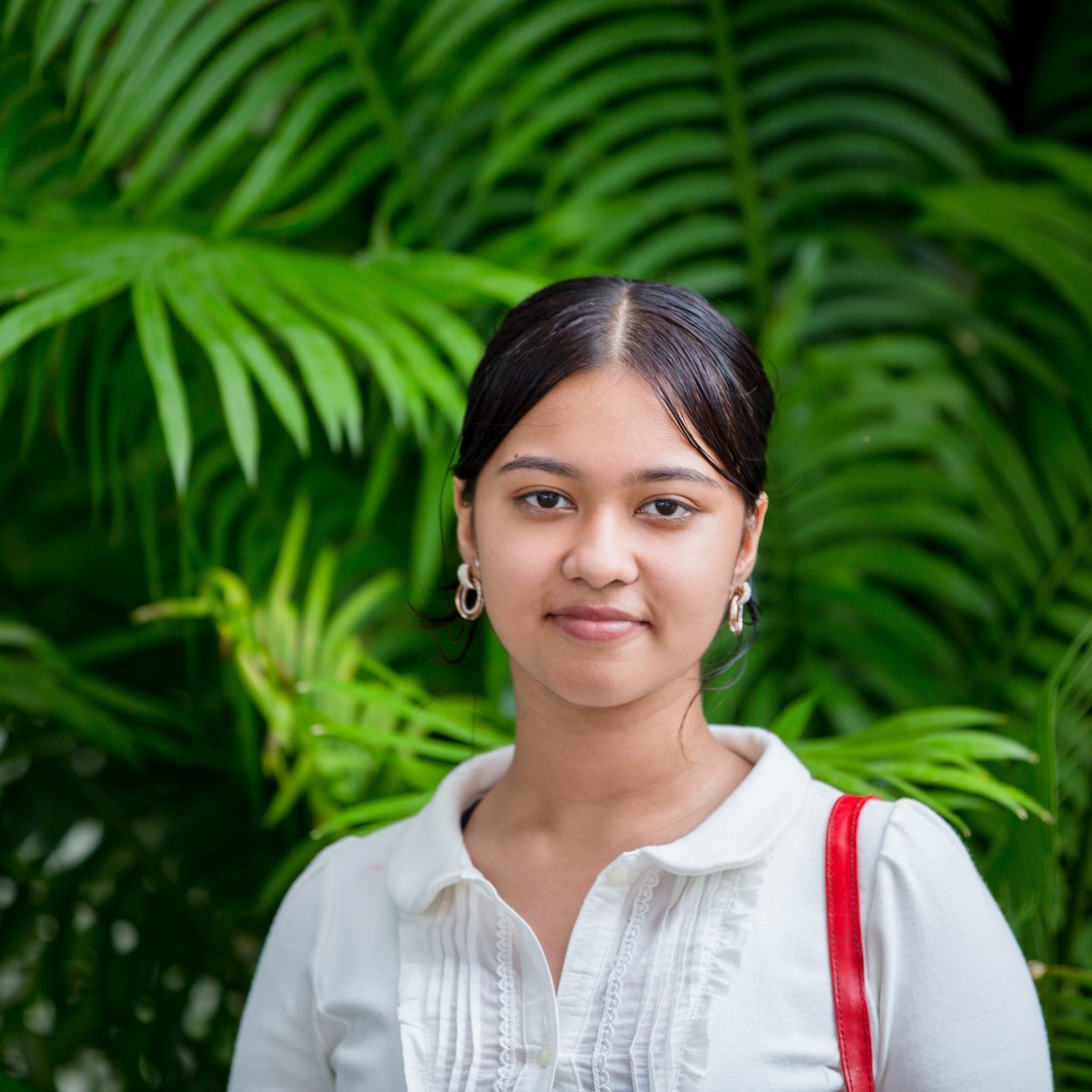 A young girl is smiling as she looks at the camera. Her is tied up and she is wearing a white shirt with a red handbag on her shoulder. She is a participant of FWRM's GIRLS program. The photo was taken during one of FWRM's GIRLS workshop.