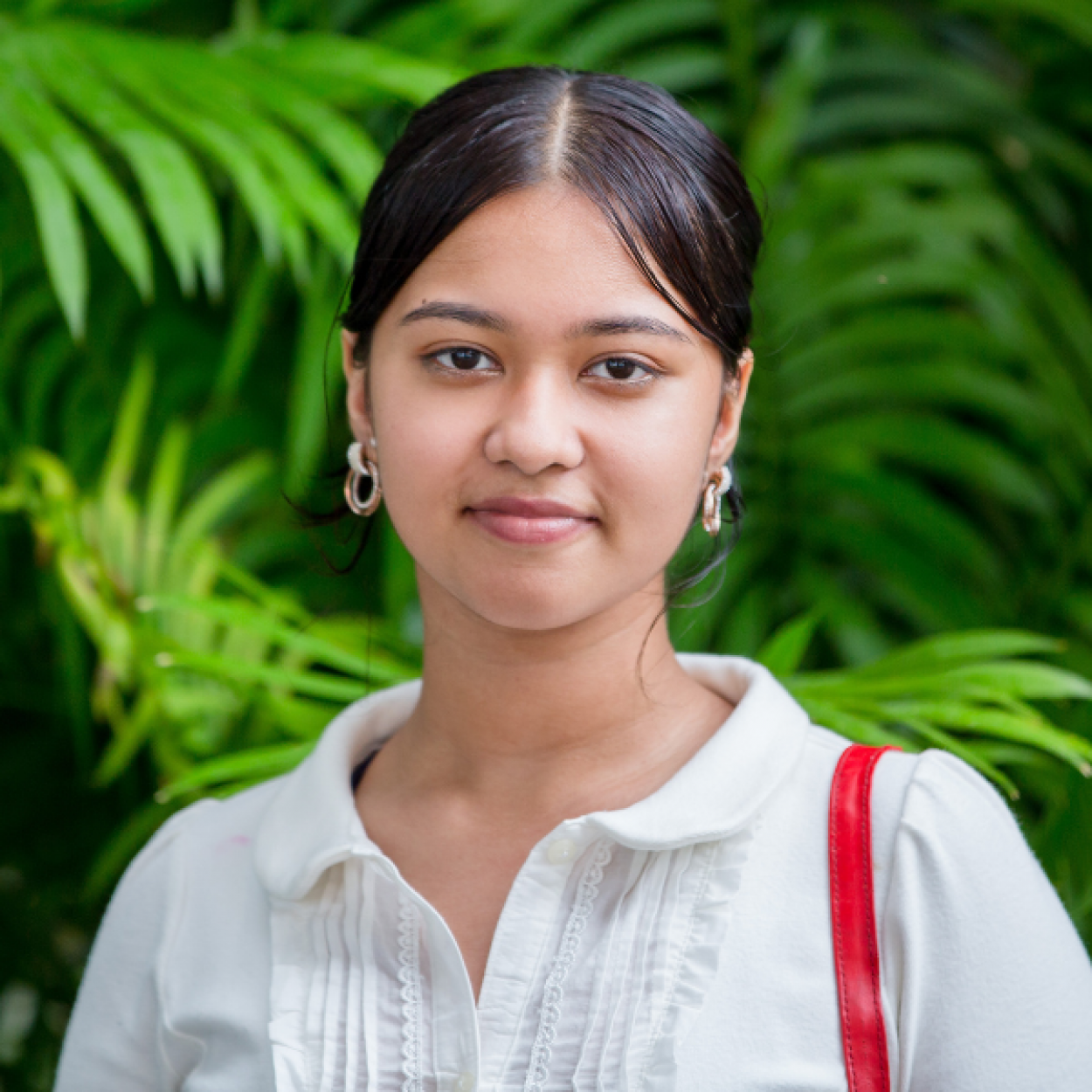 A young girl is smiling as she looks at the camera. Her is tied up and she is wearing a white shirt with a red handbag on her shoulder. She is a participant of FWRM's GIRLS program. The photo was taken during one of FWRM's GIRLS workshop.