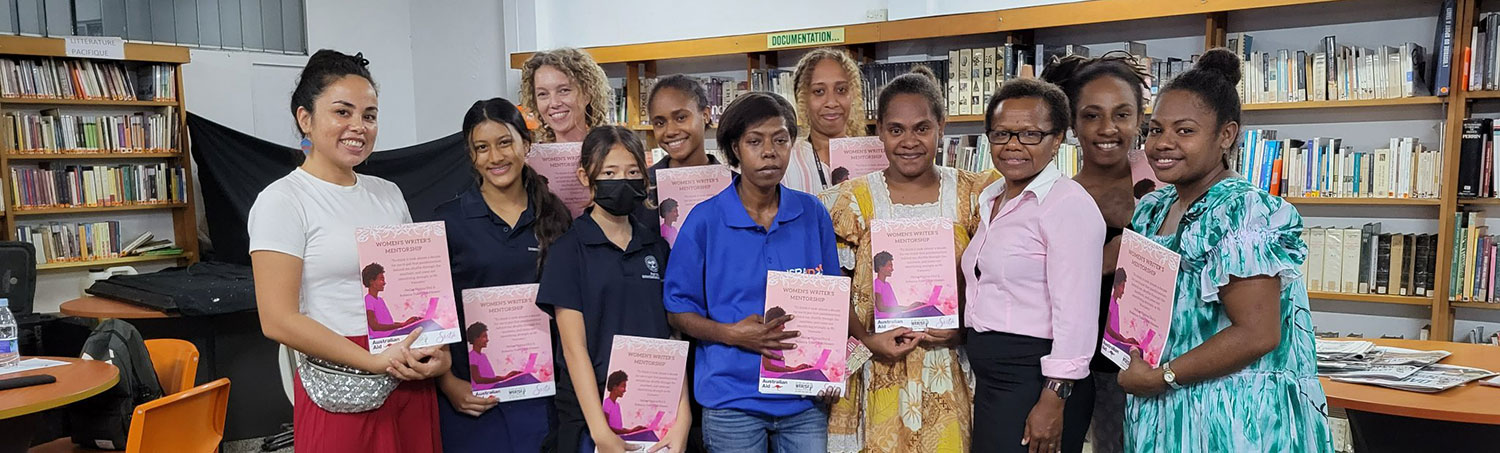 A group of women stand in a library space holding certificates from a Women Writer's Workshop