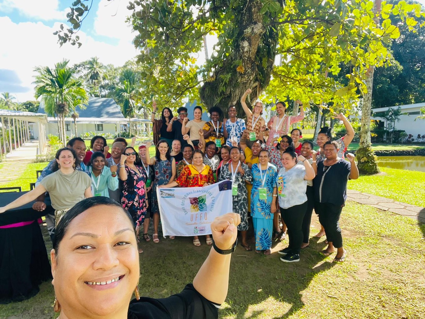 A group of people smiling and with fists raised under the branches of a large leafy tree