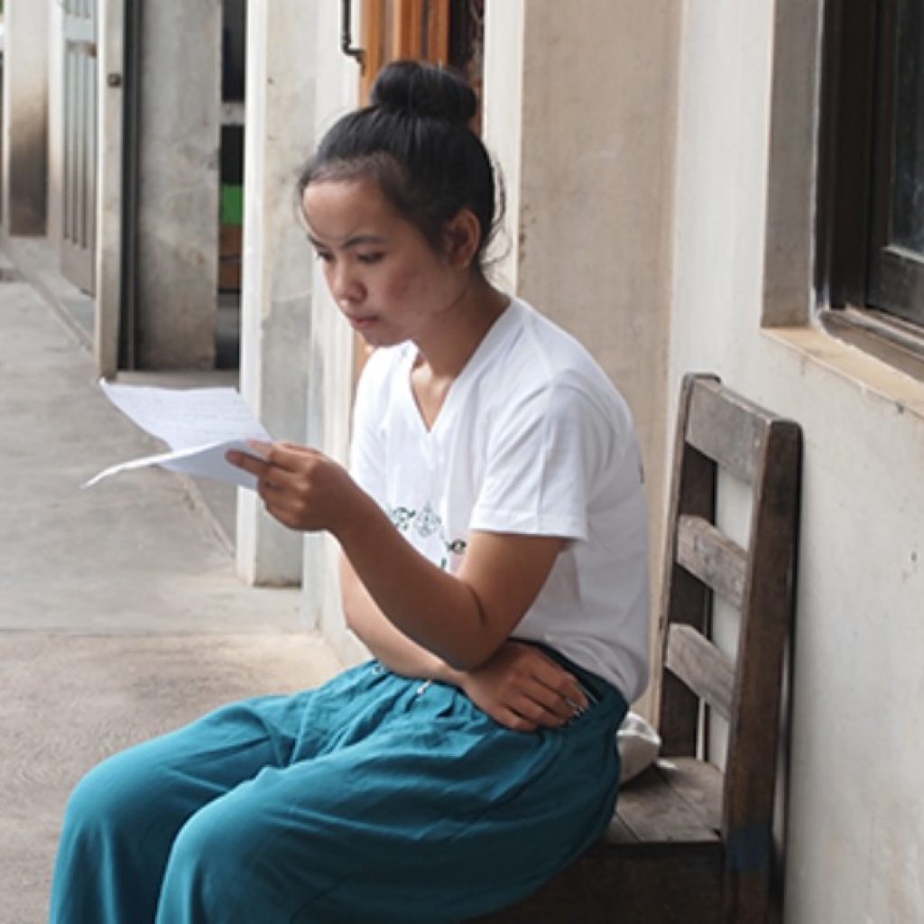 Image of a woman sitting in a hallway, reading a piece of paper.