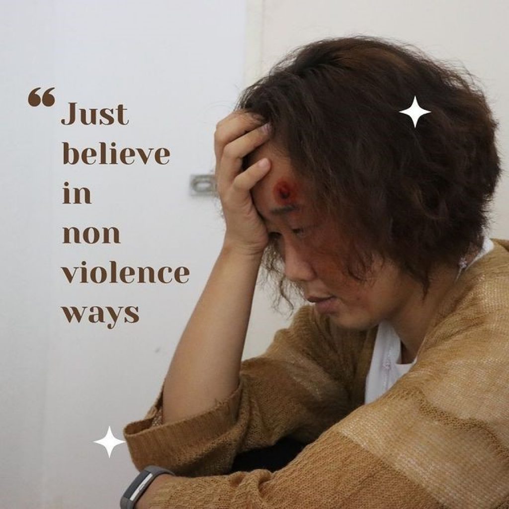 A woman with a bruised face holds her head in hands on the right of frame. The text on the left reads "just believe in non violence ways"