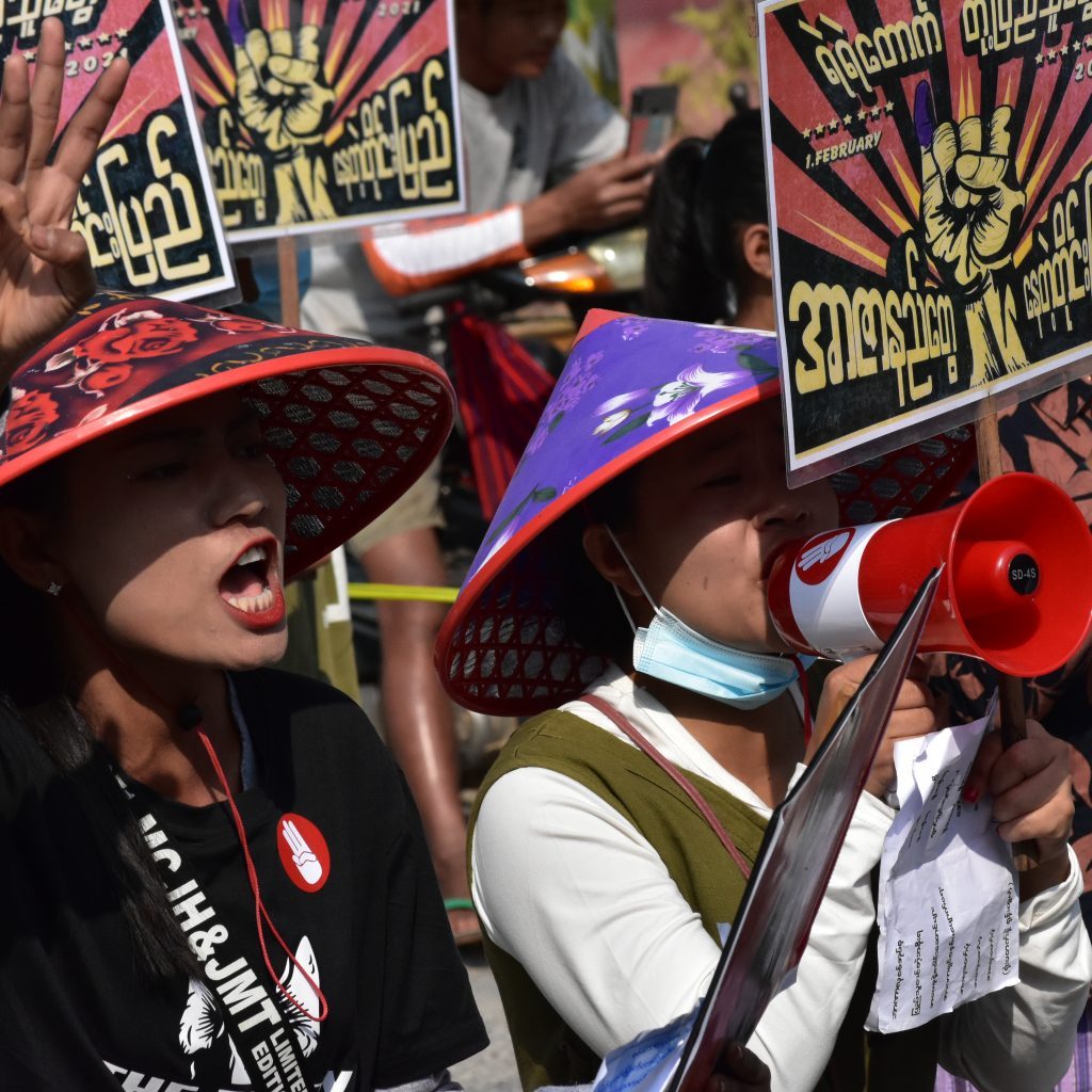 A photo of women protestors in Myanmar during a demonstration against the junta. One of the women is speaking into a megaphone and the other is holding her right hand up with three fingers lifted in sign of resistance.