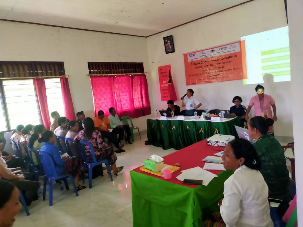 People are gathered in a room for CCC's awareness-raising session. A woman is standing at a desk set up in front of the room as she addresses the crowd through a microphone. 