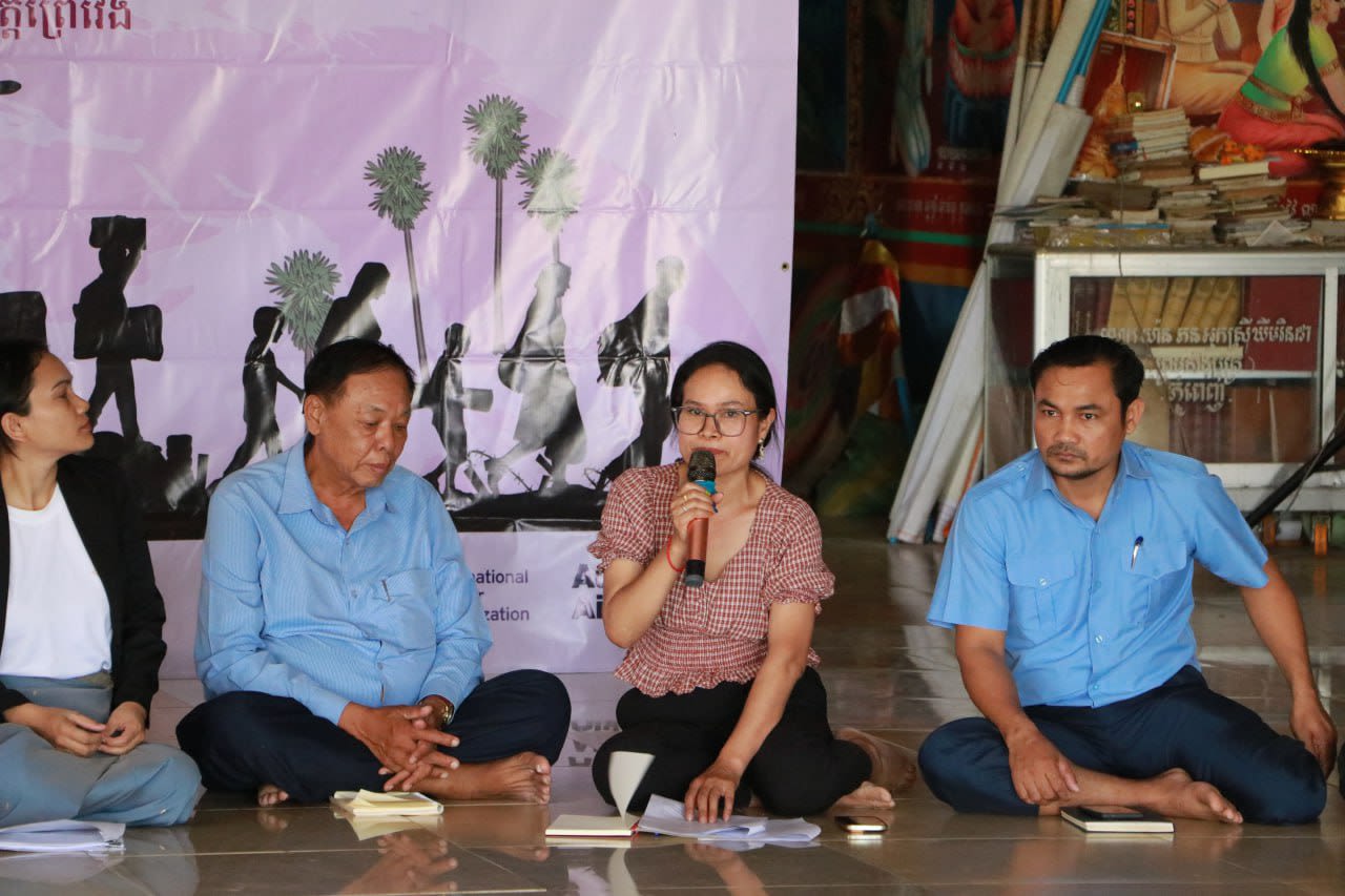 A group of men and women are seated on the floor, listening to a woman as she speaks into a microphone.
