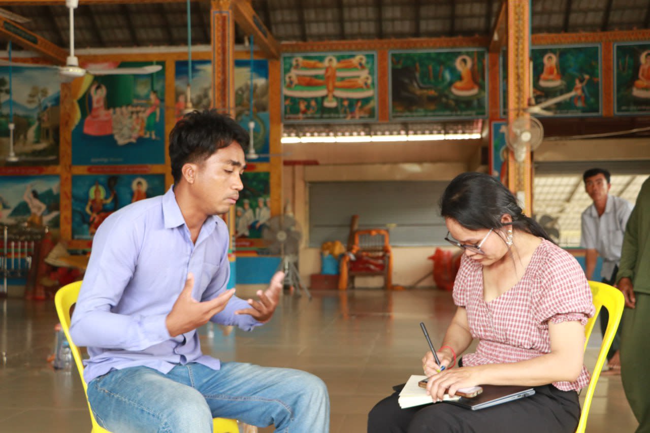 A man is havinga  conversation with a woman as she writes down notes. Both of them are seated on yellow plastic chairs.