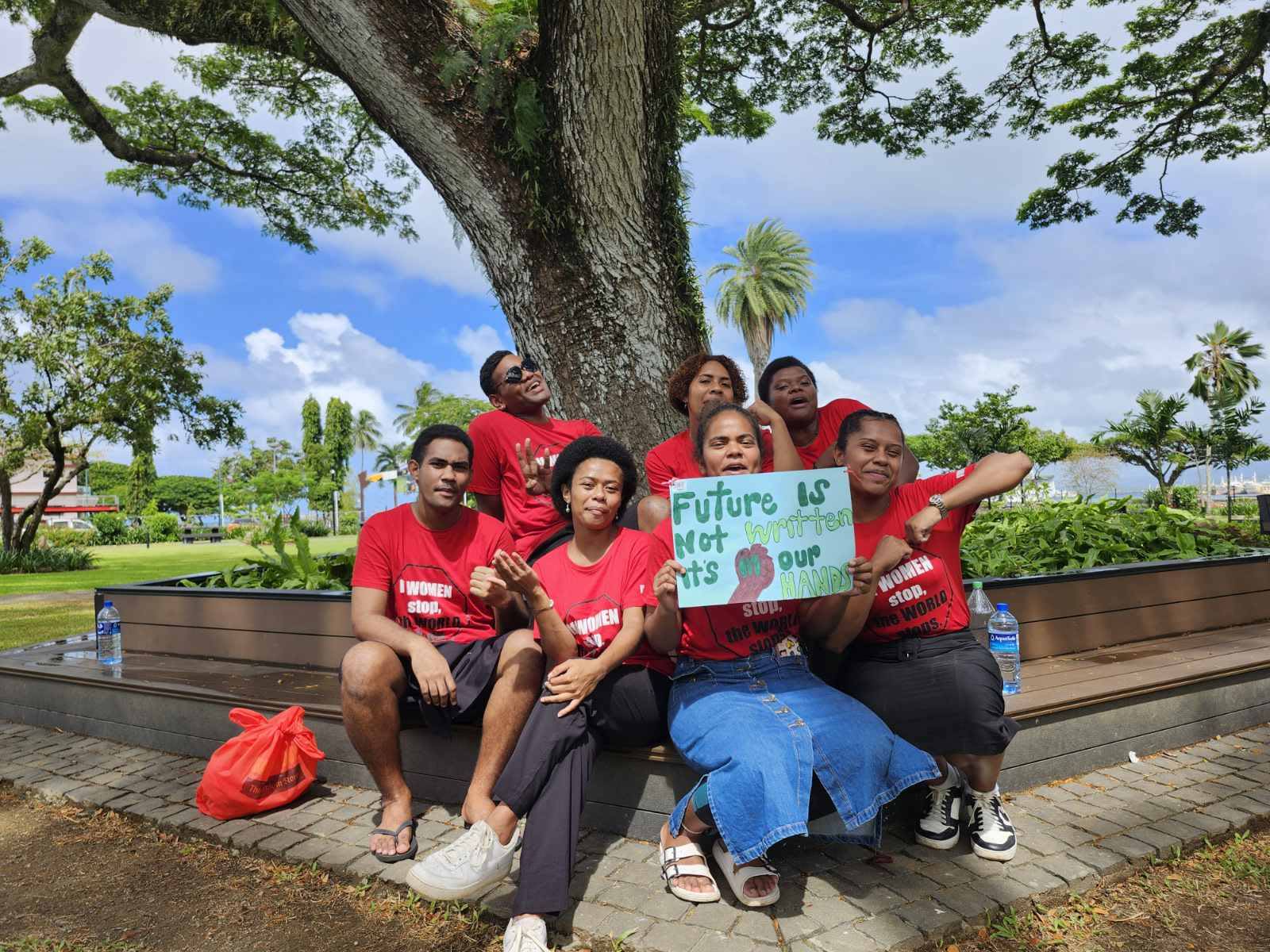 A group of men and women are seated together on a park bench at the 'If Women Stop, the World Stops' concert. They are wearing matching red concert t-shirts and chatting as they pose for the camera. One woman is holding up a sign that reads, 'Future is not written, it's in our hands.'