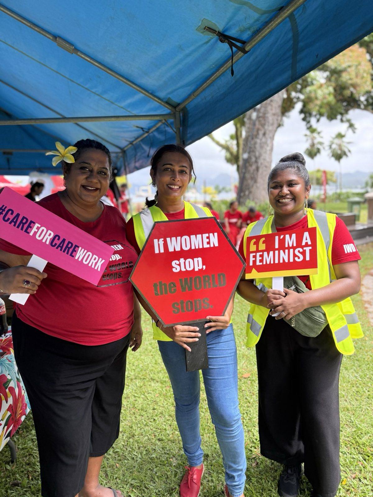 Three women, including FWRM's Nalini Singh, post together for the camera. They are smiling as they hold up signs that read, '#CareForCareWork', 'If Women Stop, the World Stops' and 'I'm a Feminist'.