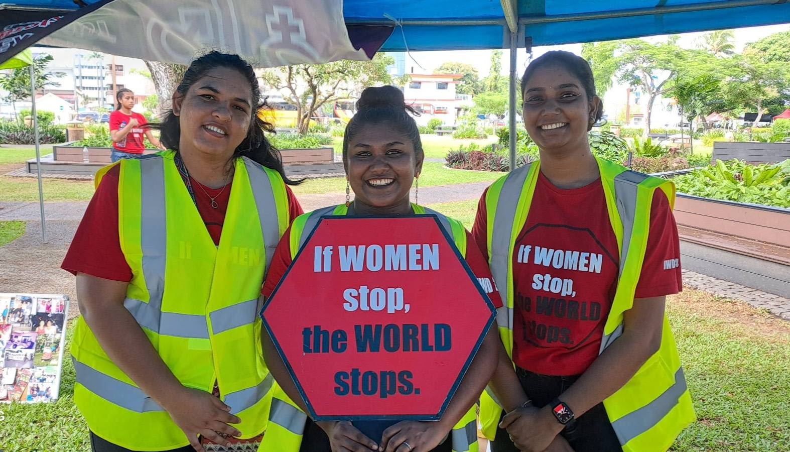 Three women post together for the camera in matching red t-shirts and hi-vis. They are smiling at the camera as one woman holds up a sign that reads, 'If Women Stop, the World Stops'. 