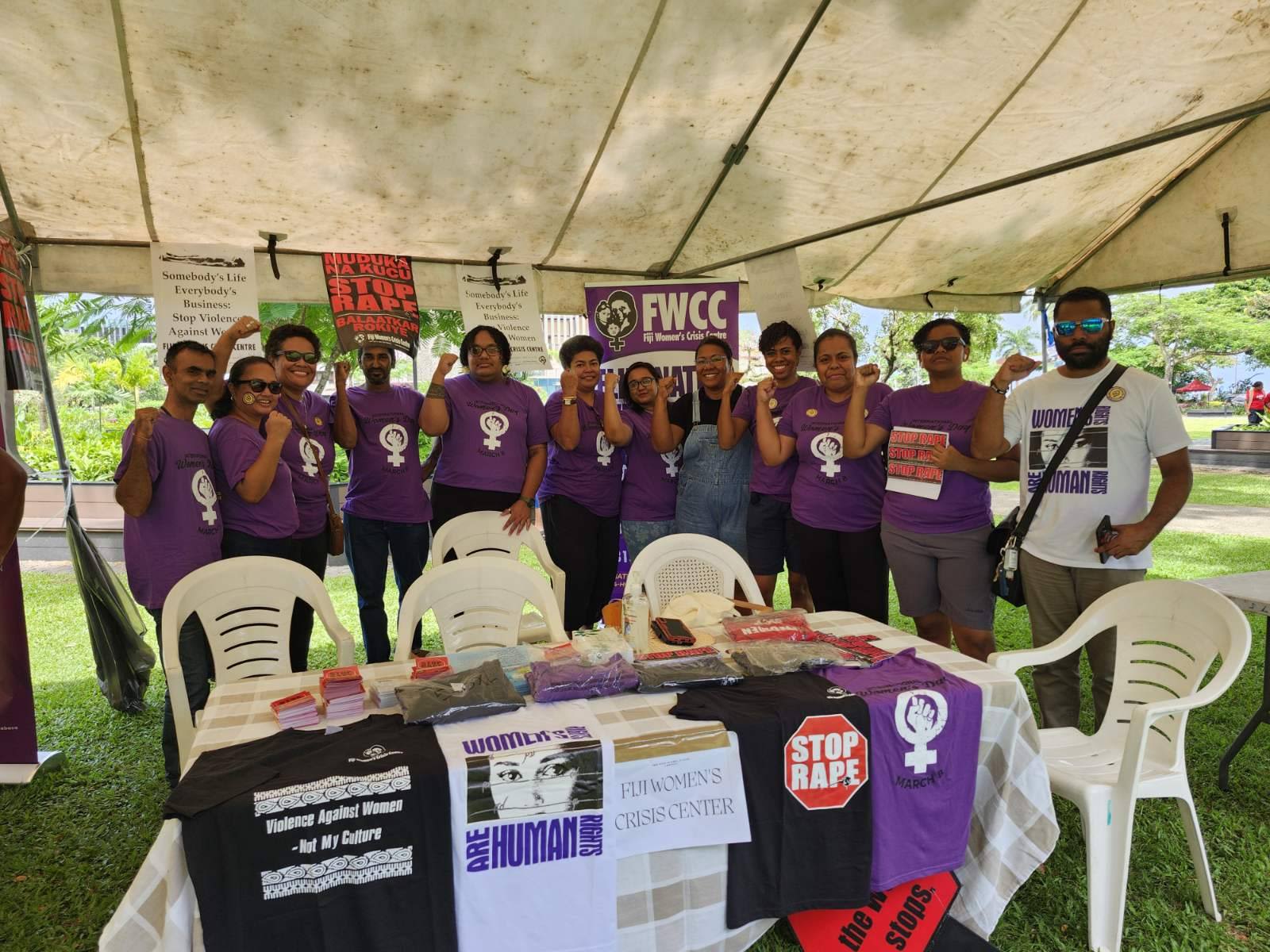 A group of staff from Fiji Women's Crisis Centre pose together at their booth. They are wearing matching purple t-shirts and holding up their fists in a symbol of strength.
