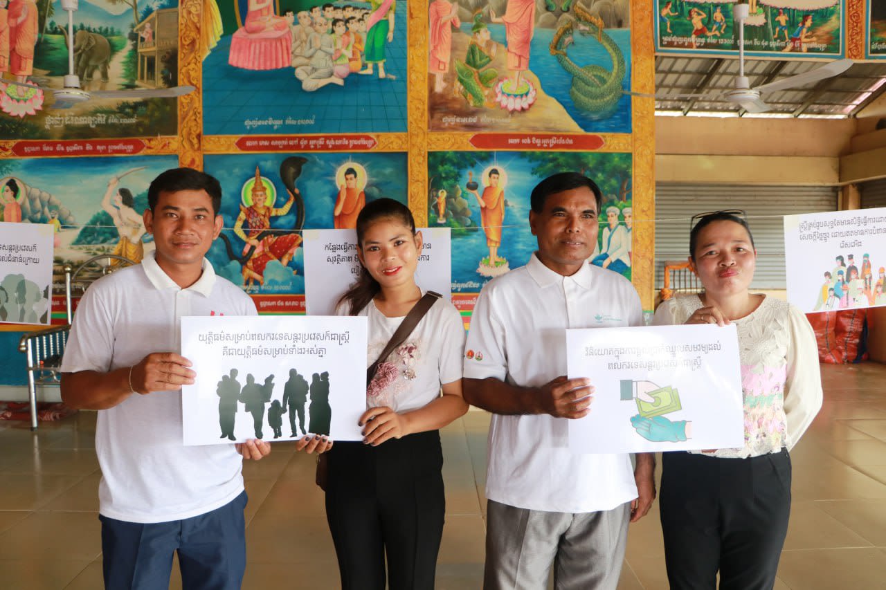 A group of men and women pose together with signs written in Khmer as they smile at the camera. 