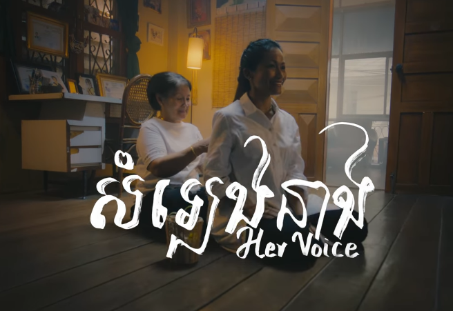 A still from the music video 'Her Voice' depicting two women seated on the floor in a house as an older woman braids the younger woman's hair.