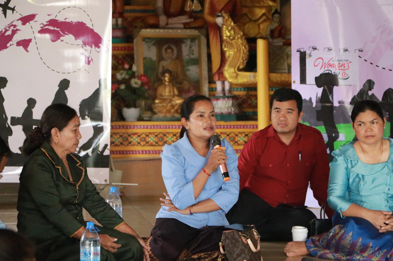 A group of men and women are seated on the floor, listening to a woman as she speaks into a microphone.