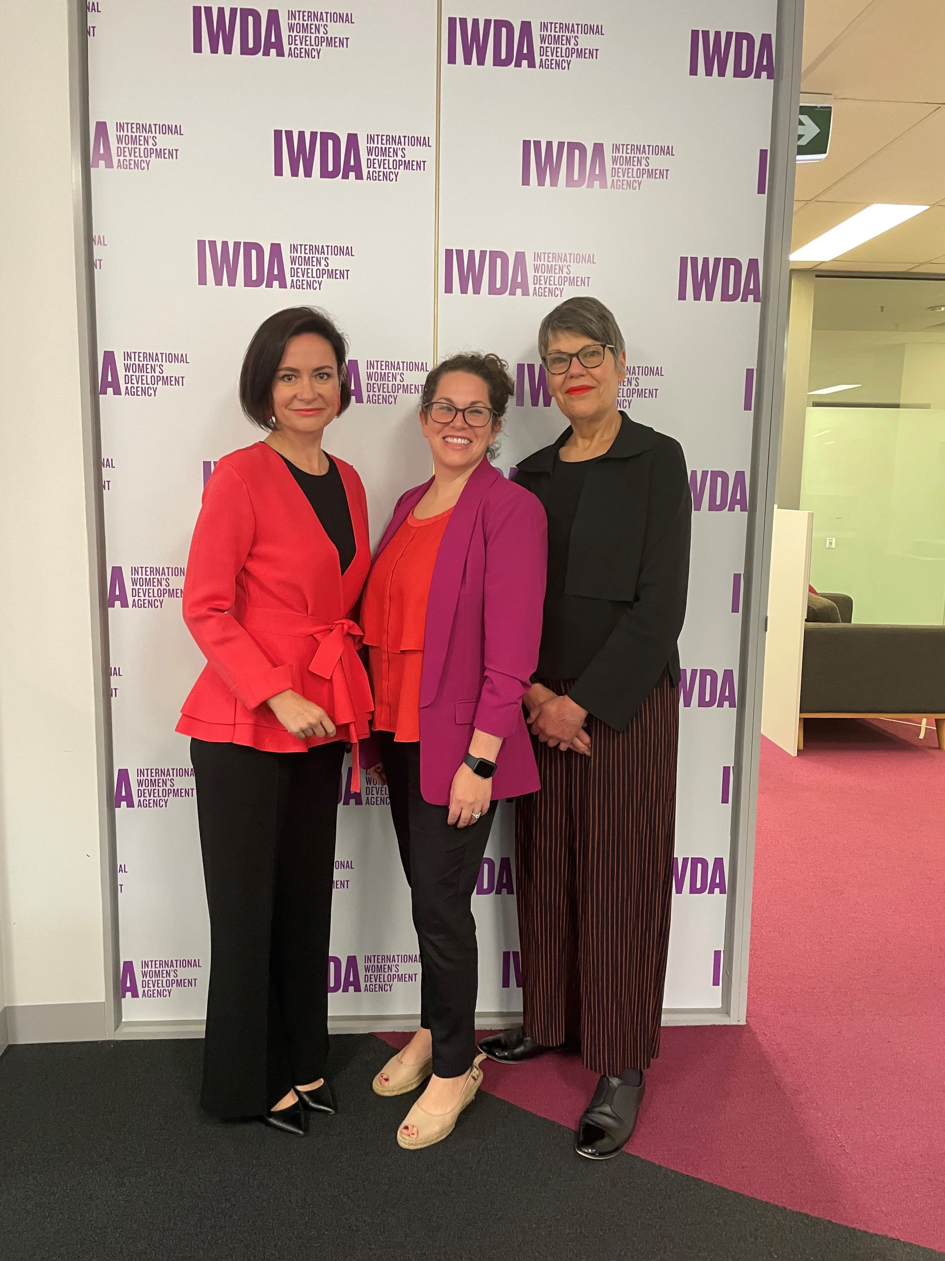 Bettina, Jo Pradela and Jo Crawford are standing together as they smile for the camera. They are standing in front of an IWDA photowall wearing the Equality Insights brand colours.
