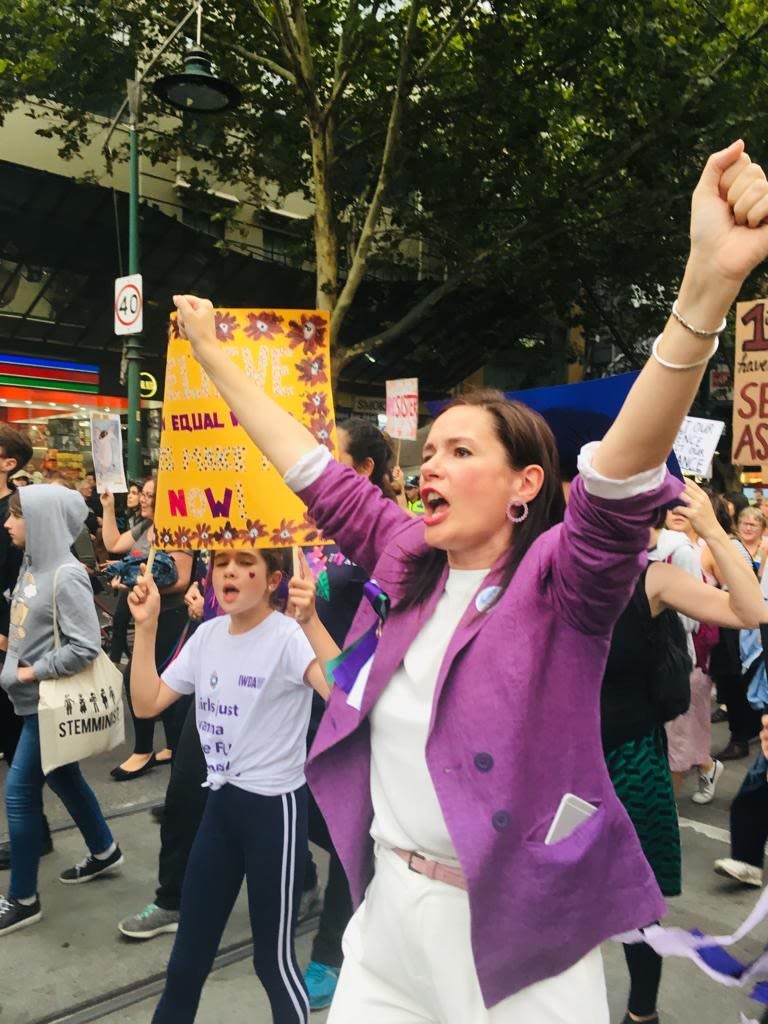 Bettina and her daughter attend an International Women's Day march. Bettina is wearing a purple blazer over a white top and has her arms raised as she chants. Her daughter is holding a homemade sign and is chanting.