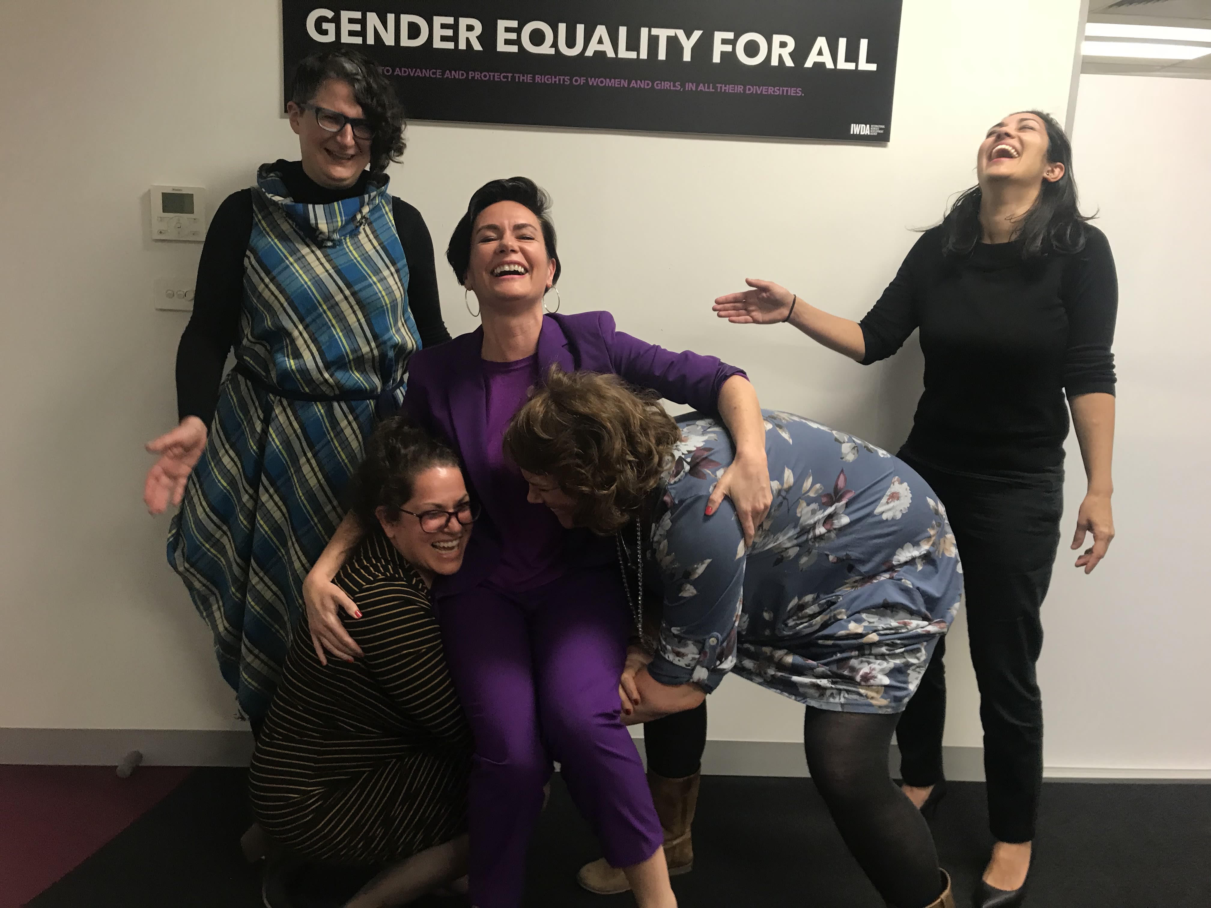 Jo and Emily are laughing as they attempt to lift a laughing Bettina up in the air. Behind them, Che and Nicky are laughing as they watch them. Bettina, always on brand, is a wearing a purple suit.