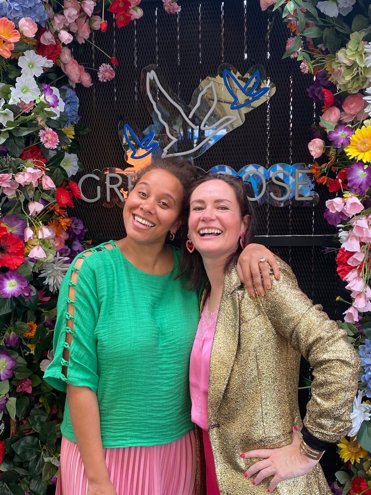 Bettina and Carla are posing together in front of a photo wall covered in flowers. They are smiling at the camera and have their arms around each other.