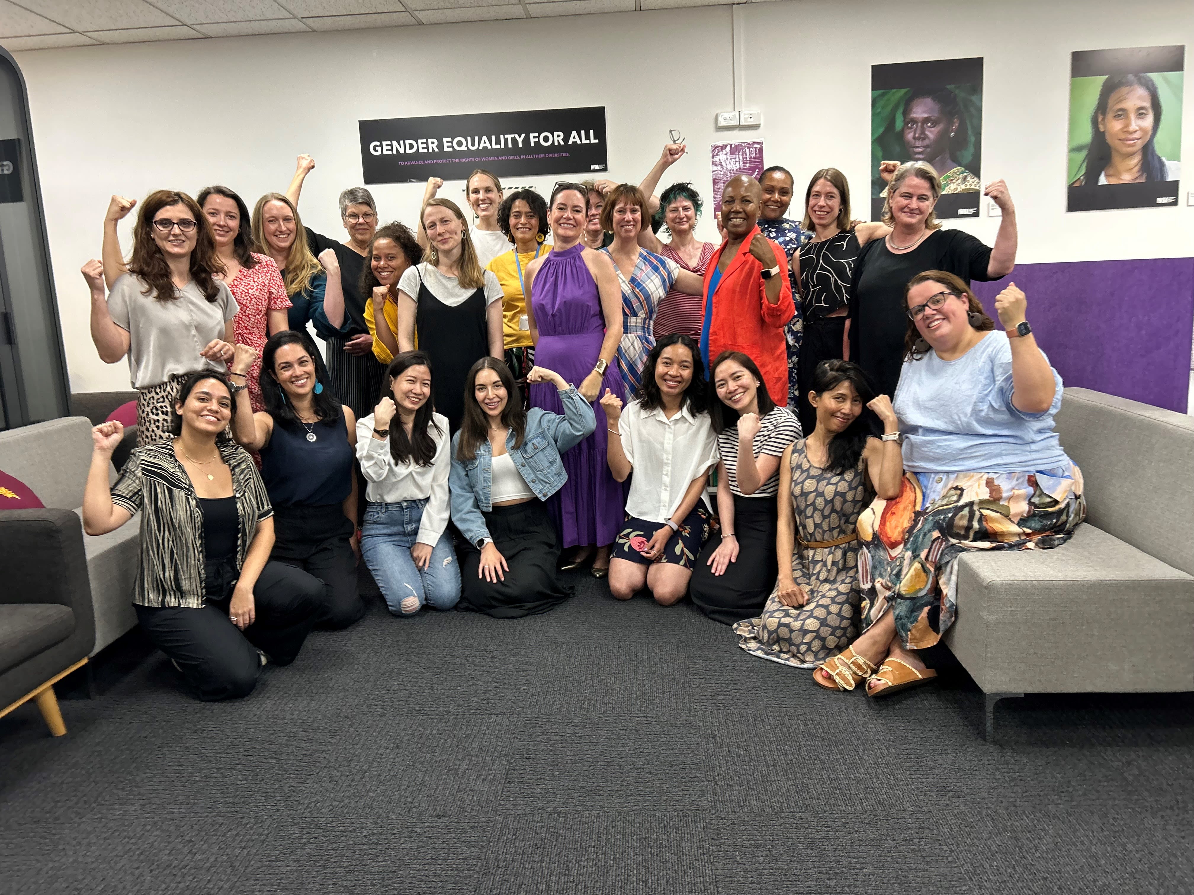 Bettina and IWDA staff members are gathered under a banner in the IWDA office that reads, "Gender equality for all." They are smiling and have their fists raised.
