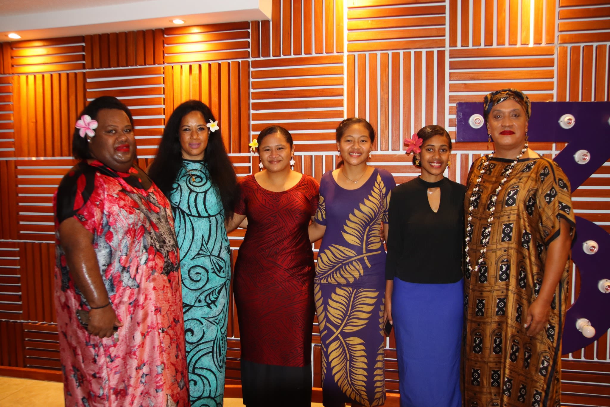 Six women smile for camera as they pose together in front of a wooden wall. 