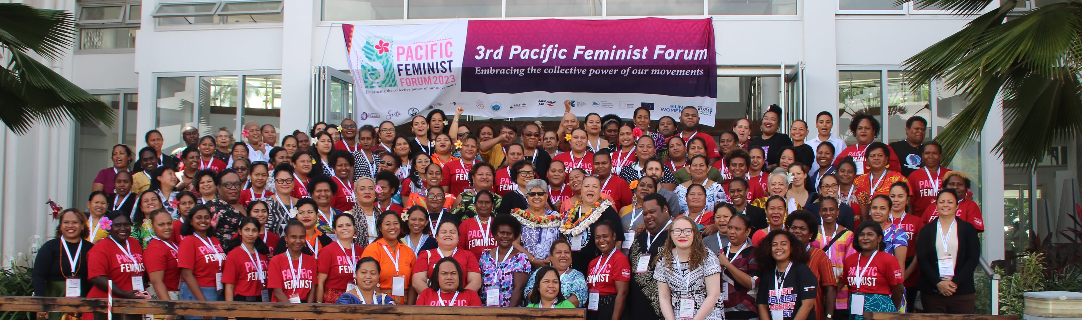 All participants at the 3rd Pacific Feminist Forum are posing together in an outdoor space in front of the event banner. Many are wearing red t-shirts that read 'Pacific Feminist'