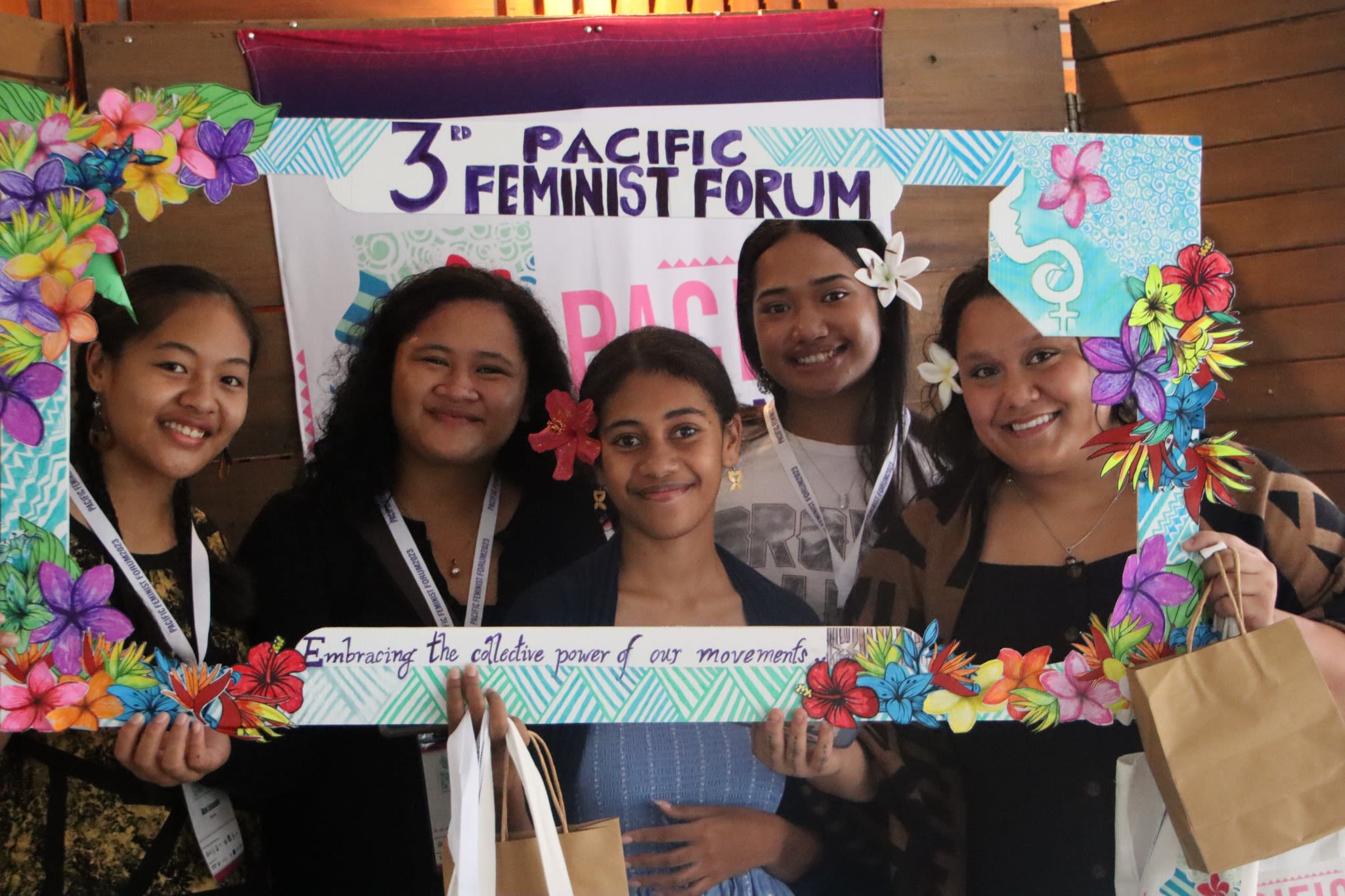 Five women are smiling at the camera as they pose with a selfie frame around which the words '3rd Pacific Feminist Forum: Embracing the collective power of movements' are written. The frame is decorated with hand-drawn flowers from the Pacific against a backdrop of water.