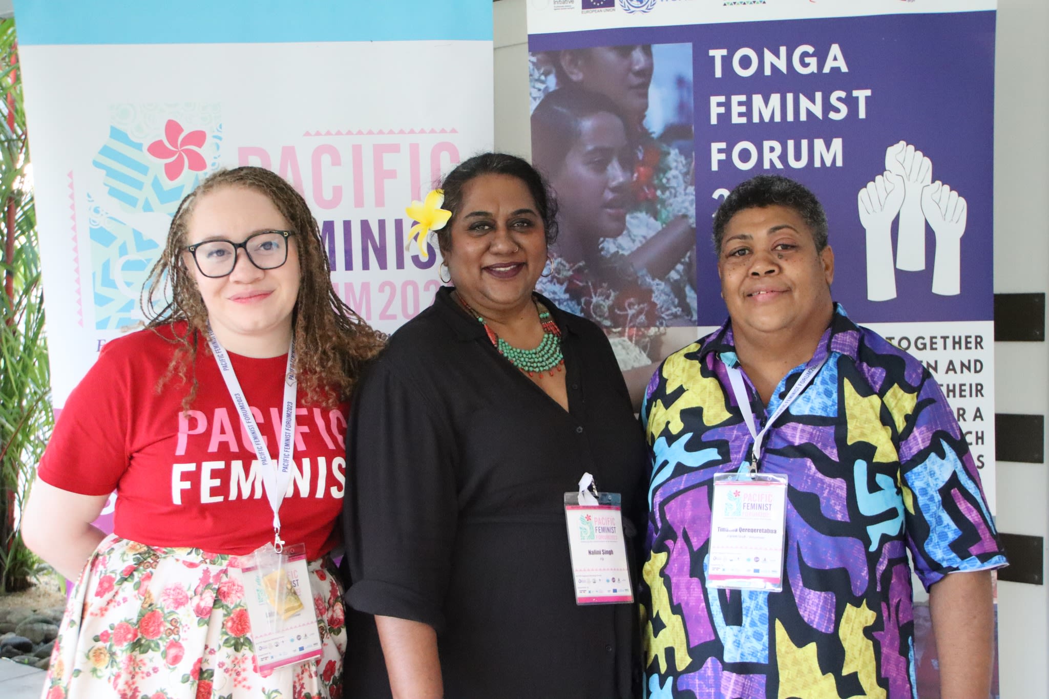 Three women are smiling at the camera as they pose in front of banners that read 'Pacific Feminist Forum' and 'Tonga Feminist Forum'.