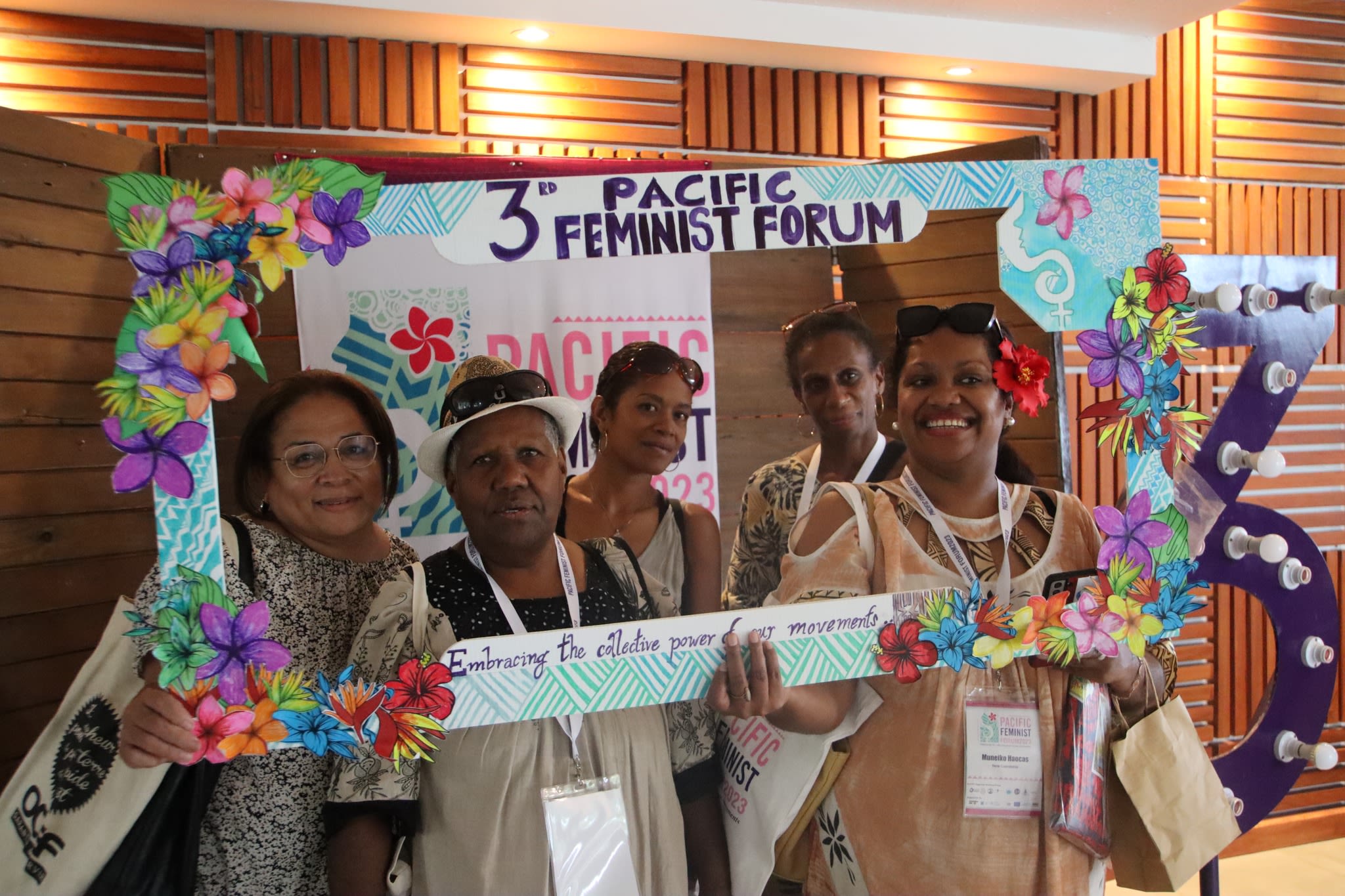Five women are smiling at the camera as they pose with a selfie frame around which the words '3rd Pacific Feminist Forum: Embracing the collective power of movements' are written. The frame is decorated with hand-drawn flowers from the Pacific against a backdrop of water.