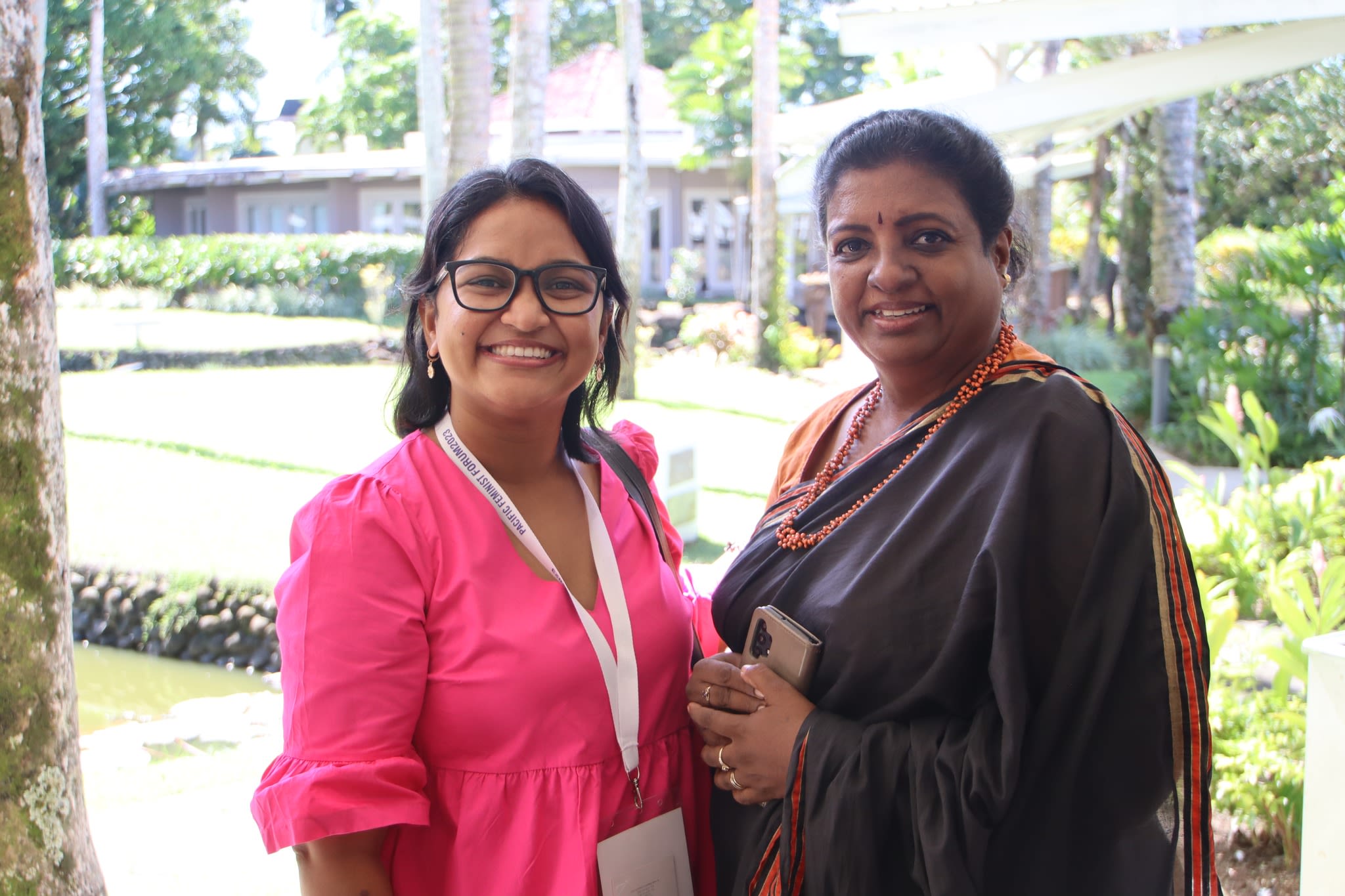 Two women are smiling at the camera as the pose together in a garden at the 3rd Pacific Feminist Forum.
