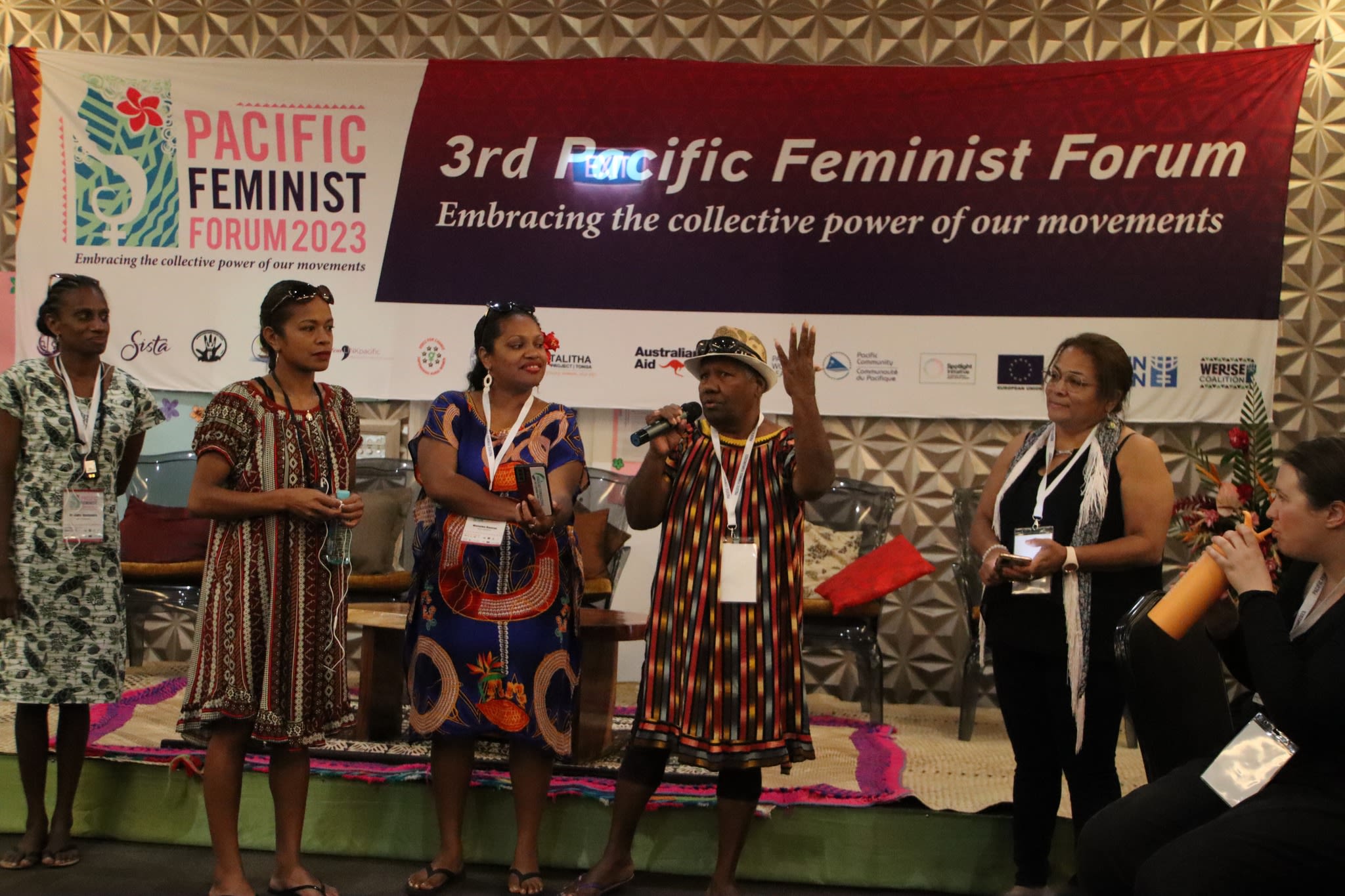 Five women are standing together on stage in front of a banner that reads '3rd Pacific Feminist Forum: embracing the collective power of our movements'. One of the women is holding a microphone as she addresses participants at the forum.