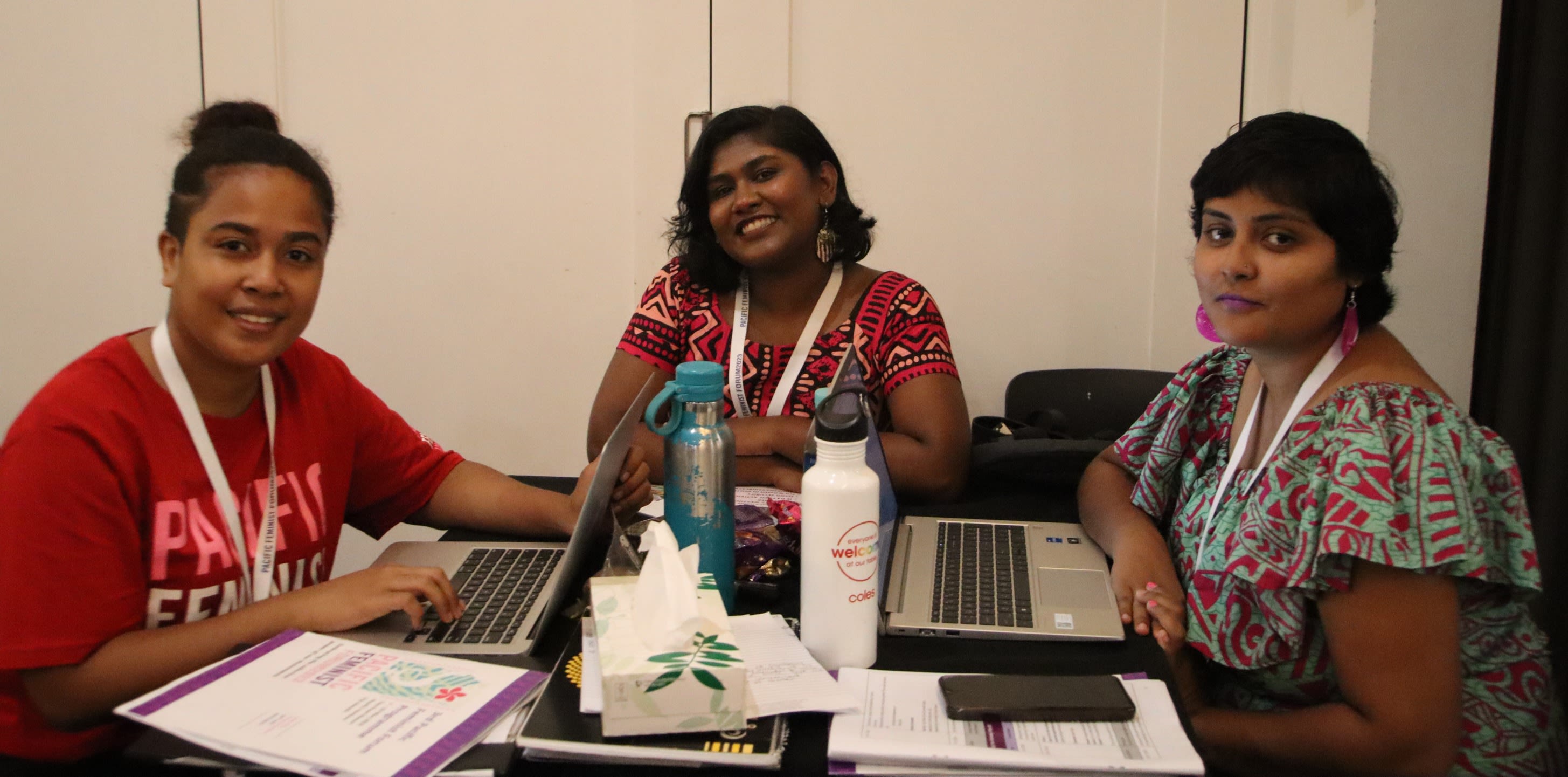 Three women are smiling for the camera. They are seated around a table with laptops and forum documents in front of them.