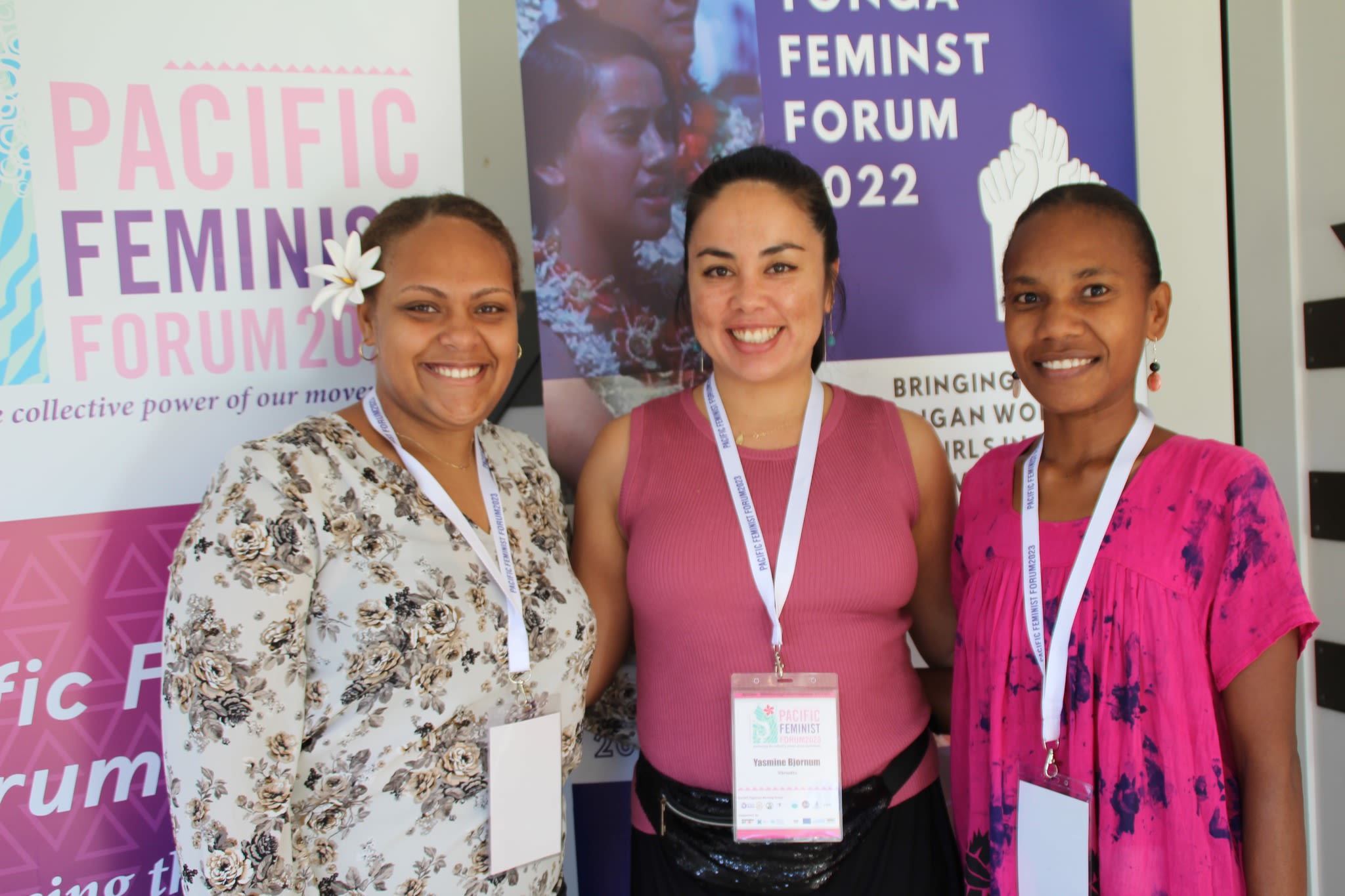 Three women are posing together for the camera. They are standing in front of banners that read 'Pacific Feminist Forum 2023' and 'Tonga Feminist Forum 2022.'