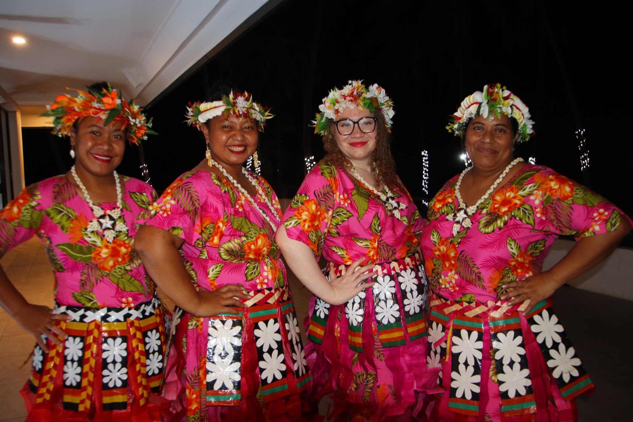 Four women are posing together with their hands on their hips. It is nighttime and they are wearing traditional Fijian dress.