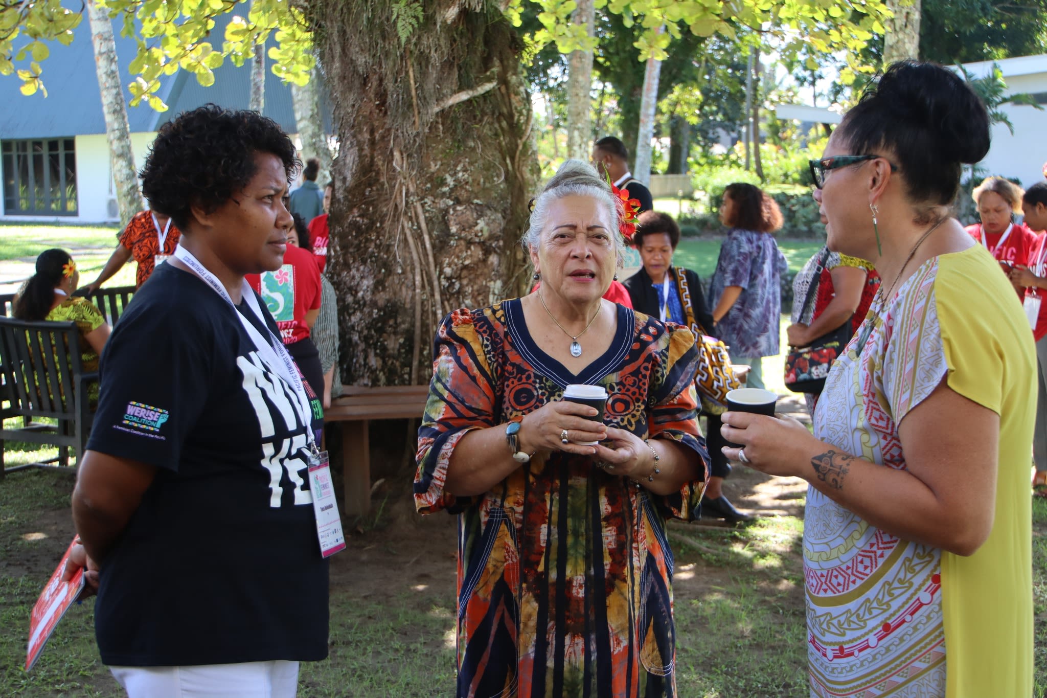 Three women are deep in conversation during a break from the conference. They are standing outside in front of a large tree with groups of people behind them.