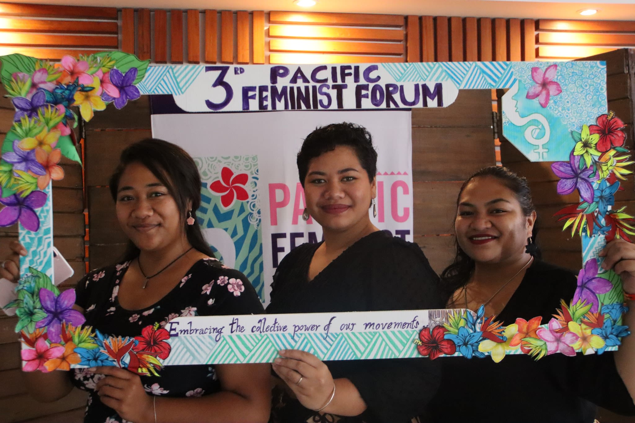 Three women are smiling at the camera as they pose with a selfie frame around which the words '3rd Pacific Feminist Forum: Embracing the collective power of movements' are written.