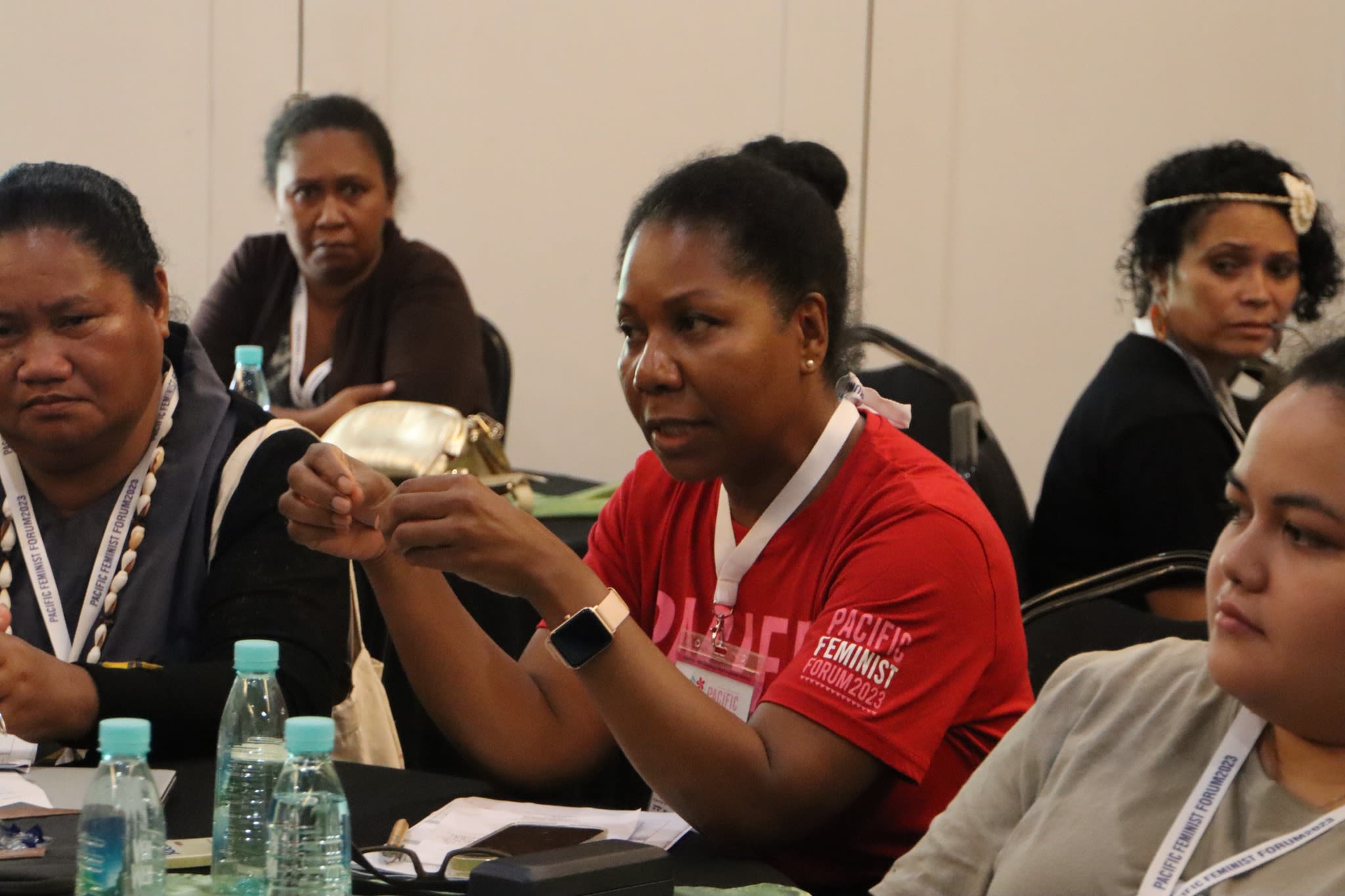 A woman is speaking with others around a table. She is wearing a red t-shirt that reads 'Pacific Feminist Forum 2023.'