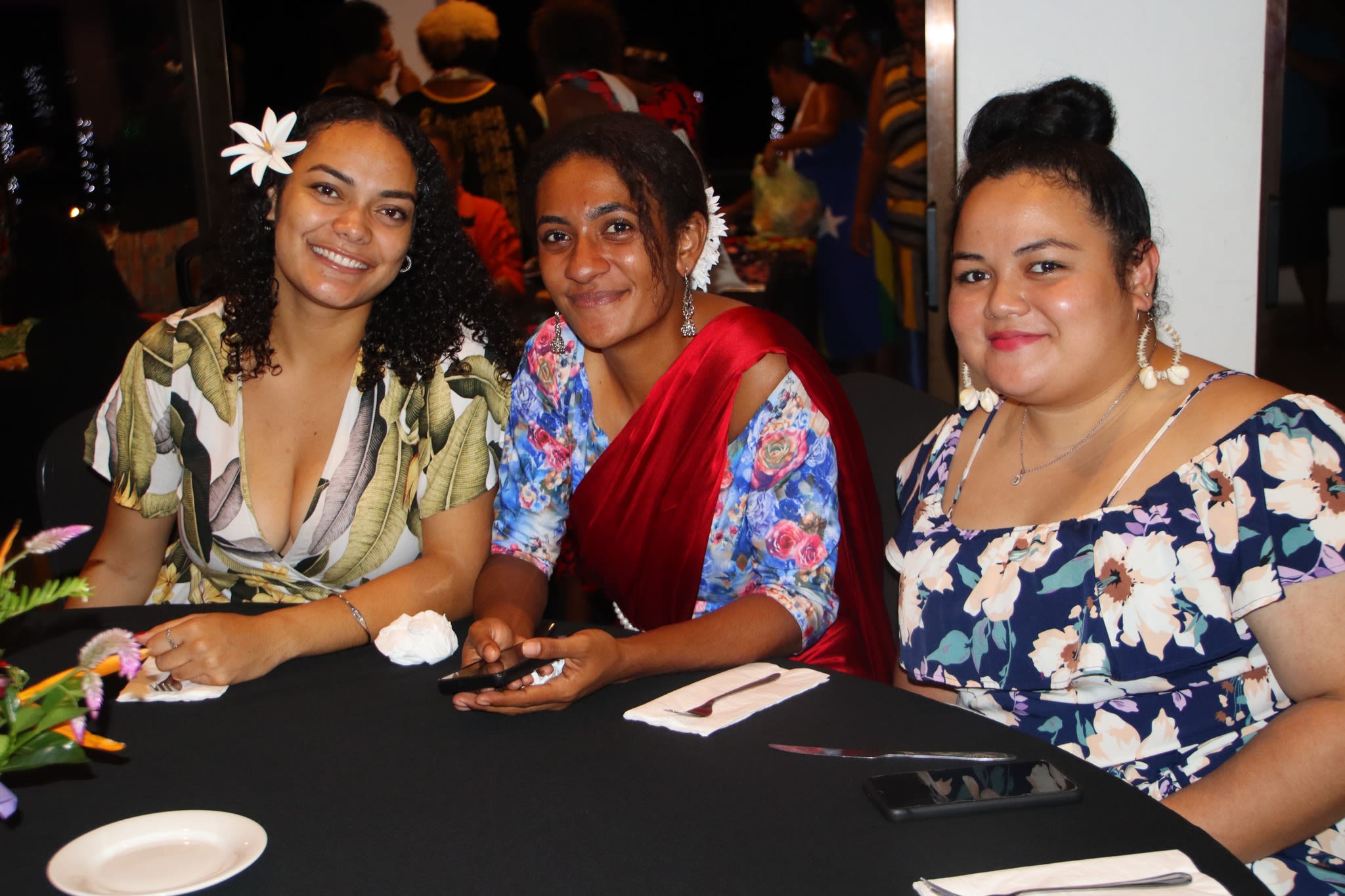 Three women are seated together at a table and smiling at the camera.