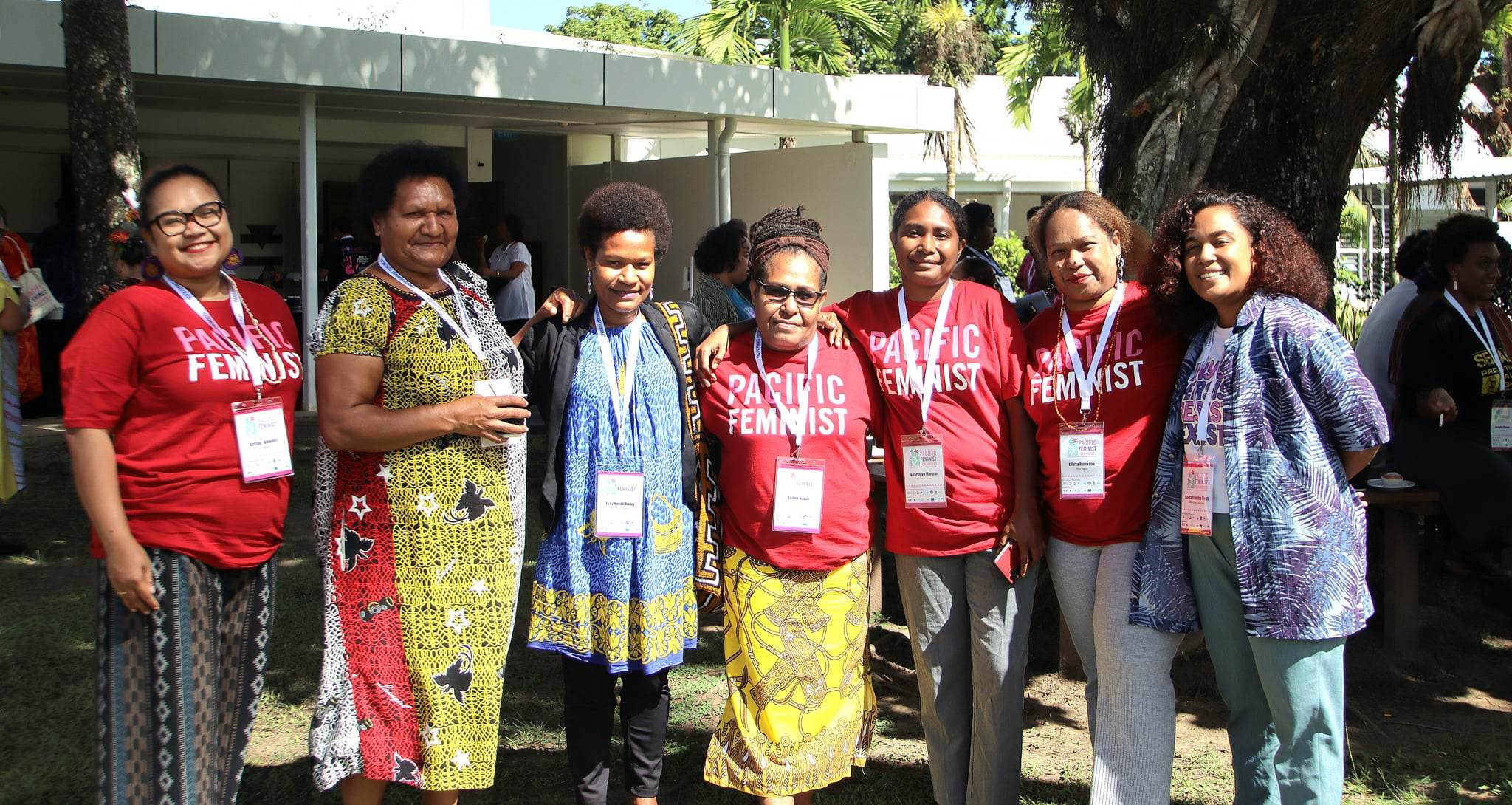 Seven women are standing together in an outdoor area as they pose for the camera. Some of them are wearing red t-shirts that read 'Pacific Feminist.'