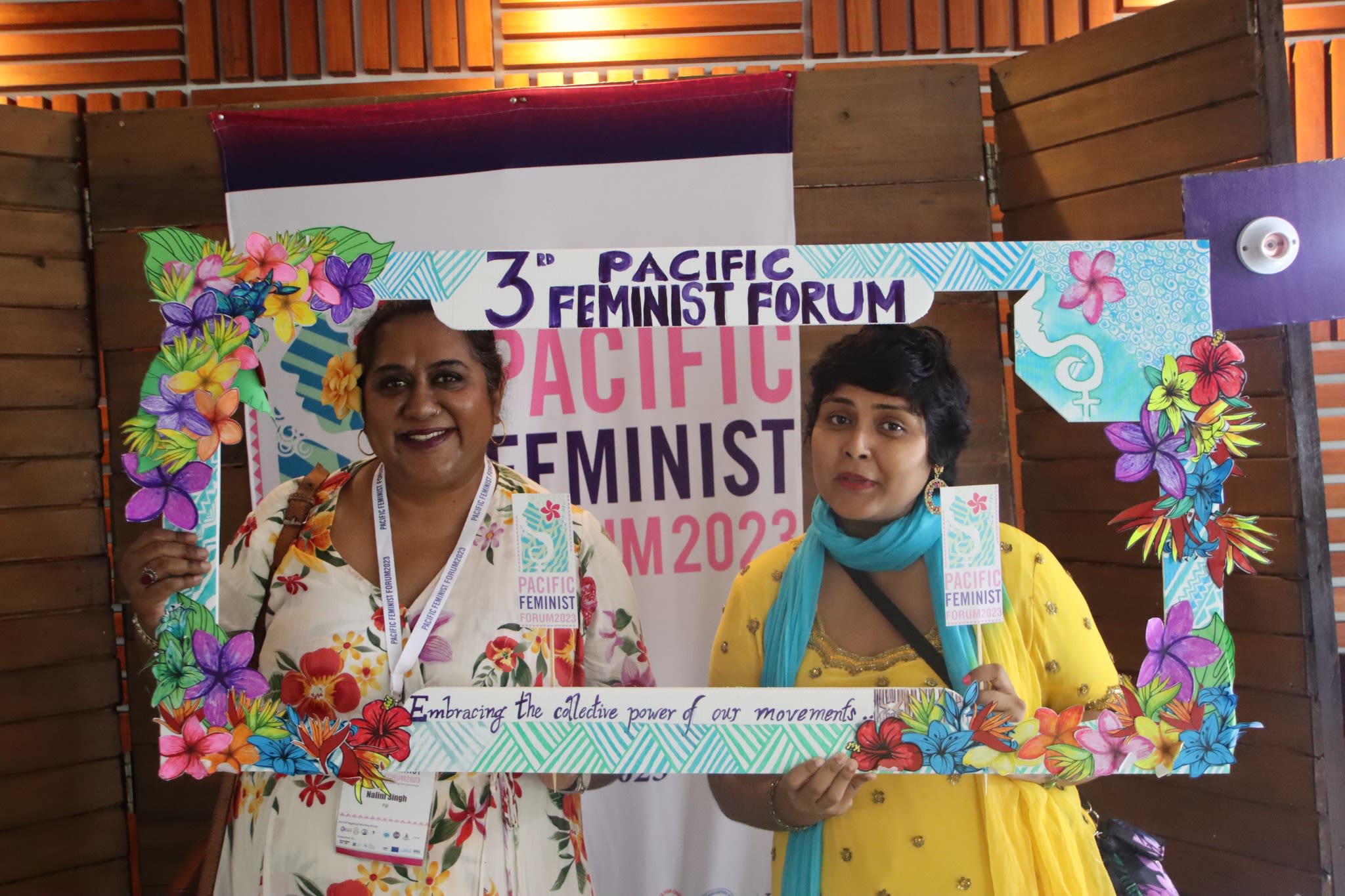 Two women are smiling at the camera as they pose with a selfie frame around which the words '3rd Pacific Feminist Forum: Embracing the collective power of movements' are written. The frame is decorated with hand-drawn flowers from the Pacific against a backdrop of water.