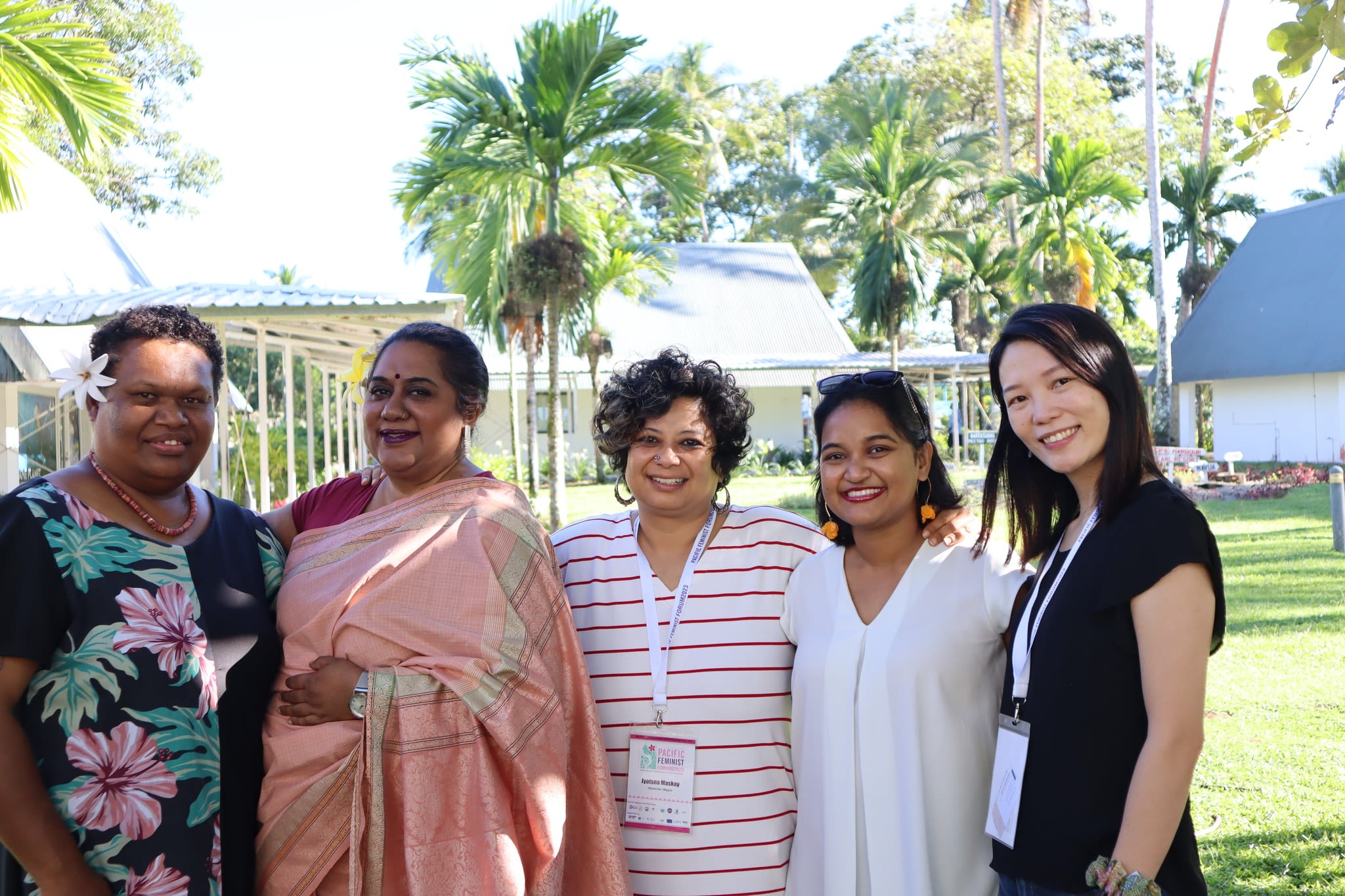 Five women are smiling at the camera as they pose together in a garden.